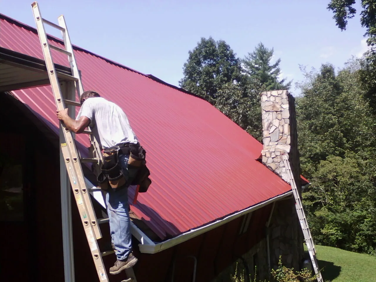 Roofer on a ladder beside a red metal roof and stone chimney — Soffit & Fascia Repair by Roofing Brothers in Fennville